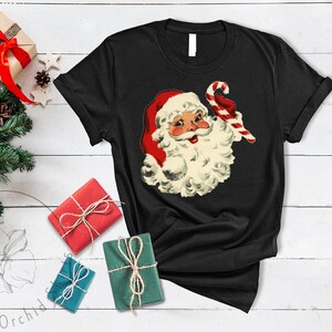 May include: Black t-shirt with a vintage illustration of Santa Claus holding a candy cane. The illustration is in red, white, and green.