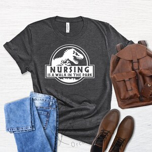 May include: A dark gray t-shirt with a white graphic of a dinosaur and the text "Nursing is a walk in the park".