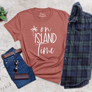 May include: A rust-colored t-shirt with the text "on island time" in white lettering. The shirt is laid flat on a gray surface with a pair of blue jeans, a brown leather belt, sunglasses, and a brown leather case.