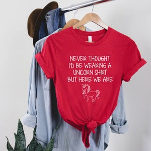 May include: Red t-shirt with a white graphic that says "Never thought I'd be wearing a unicorn shirt but here we are" and a white unicorn illustration.