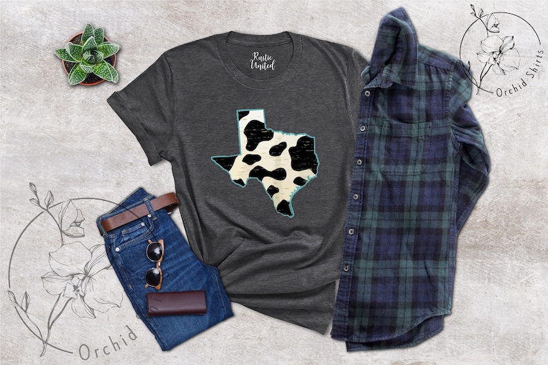 May include: A dark gray t-shirt with a white and black cow print outline of the state of Texas. The shirt is laid flat on a light gray textured surface with a pair of blue jeans, a brown leather belt, sunglasses, and a brown leather wallet.