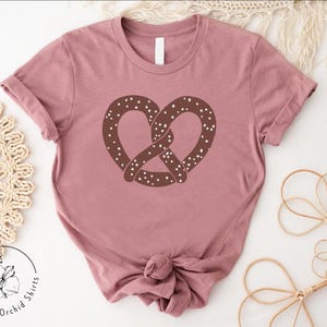 Pu&ograve; includere: Una maglietta color malva con un grafico di un pretzel marrone. Il pretzel ha delle spolverate bianche.