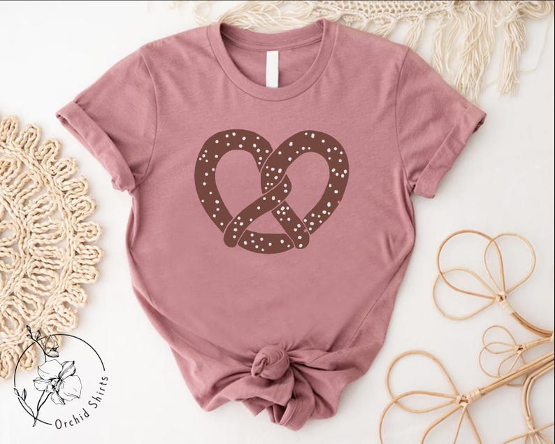 Pu&ograve; includere: Una maglietta color malva con un grafico di un pretzel marrone. Il pretzel ha delle spolverate bianche.