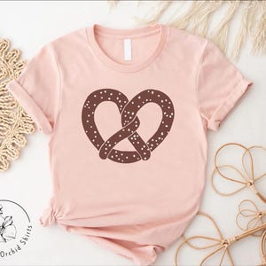 Pu&ograve; includere: Una maglietta rosa con un disegno di pretzel marrone. Il pretzel ha un design a spolverata.