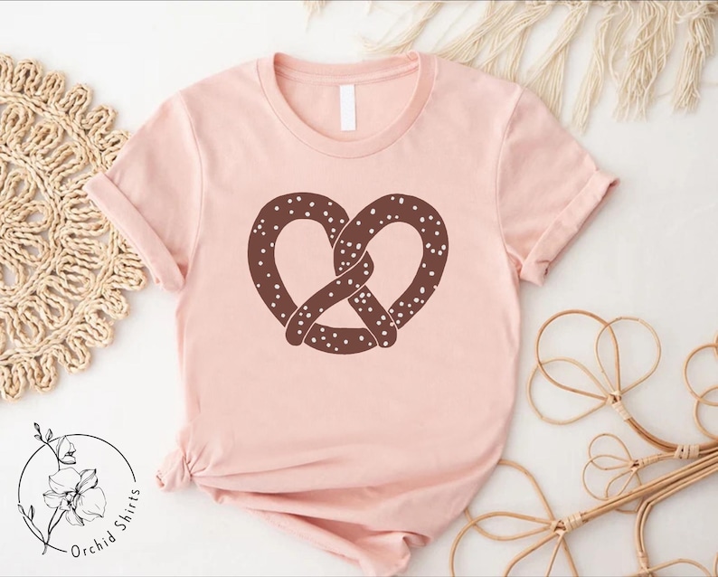 Pu&ograve; includere: Una maglietta rosa con un disegno di pretzel marrone. Il pretzel ha un design a spolverata.