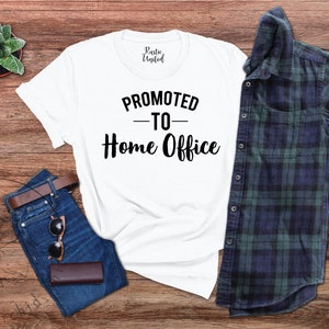 May include: A white t-shirt with the text "PROMOTED - TO - Home Office" in black. The shirt is displayed on a wooden surface alongside blue jeans, a brown belt, sunglasses, and a plaid shirt. A small succulent plant is also present.