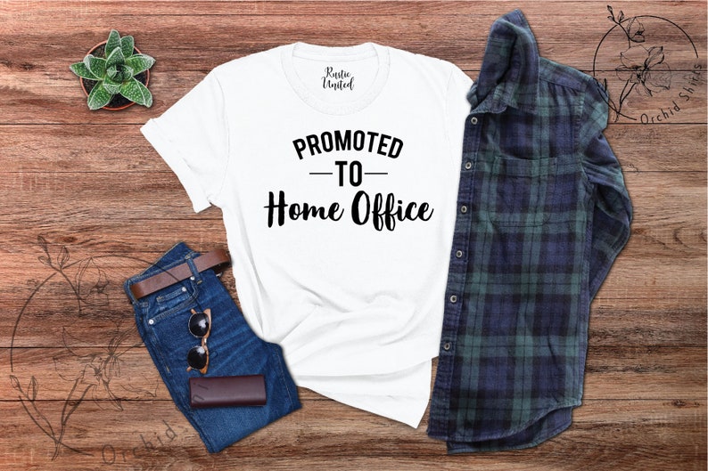 May include: A white t-shirt with the text "PROMOTED - TO - Home Office" in black. The shirt is displayed on a wooden surface alongside blue jeans, a brown belt, sunglasses, and a plaid shirt. A small succulent plant is also present.