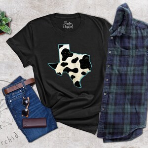 May include: Black t-shirt with a white and black cow print design of the state of Texas. The shirt is laid flat on a gray surface with a pair of jeans, a potted plant, and a plaid shirt.