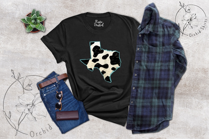May include: Black t-shirt with a white and black cow print design of the state of Texas. The shirt is laid flat on a gray surface with a pair of jeans, a potted plant, and a plaid shirt.