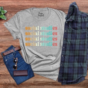 May include: A heather gray t-shirt with the words "Social Studies" in a retro font. The shirt is paired with blue jeans, sunglasses, a brown wallet, and a plaid shirt. A small succulent plant in a pot is also visible.
