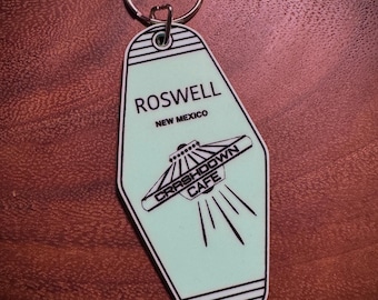 Peut inclure: Un porte-clés de style motel vert menthe avec un anneau argenté. Le porte-clés présente le texte "ROSWELL NEW MEXICO" et une illustration d'OVNI avec les mots "CRASHDOWN CAFE".