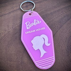 Puede incluir: Un llavero rosa con las palabras "Barbie DREAM HOUSE" y una silueta del perfil de Barbie. El llavero tiene un anillo plateado adjunto. El fondo es una superficie de madera oscura.