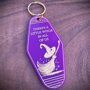 May include: A purple keychain with a gold ring. The keychain has the text "THERE'S A LITTLE WITCH IN ALL OF US" and a witch on a broom illustration in gold. The keychain is a retro hotel key style.