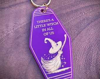 May include: A purple keychain with a gold ring. The keychain has the text "THERE'S A LITTLE WITCH IN ALL OF US" and a witch on a broom illustration in gold. The keychain is a retro hotel key style.