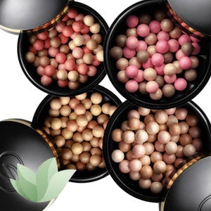 Puede incluir: Cuatro recipientes negros redondos llenos de perlas de polvo cosmético multicolor. El recipiente superior izquierdo contiene esferas rosas y bronceadas, el superior derecho esferas rosa oscuro y marrones, el inferior izquierdo doradas y beige, y el inferior derecho esferas crema y melocotón.