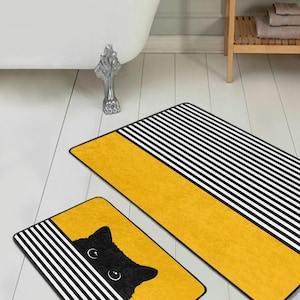 May include: Two rectangular bath mats with a playful cat design. The mats have a yellow centre with black and white striped borders. One mat features a black cat peeking out from behind the stripes.