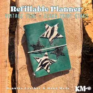 May include: A teal refillable planner featuring four star-shaped appliqués with a black and white zebra print pattern. The planner is secured with a thin white elastic band that wraps horizontally around the centre of the cover.