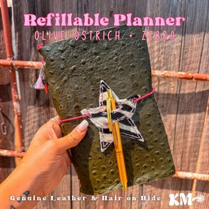 May include: A refillable planner with an olive green ostrich-textured cover, featuring a black and white zebra-print star applique. A gold-tone pen is held by the star, and a bright pink elastic band secures the planner, which has a small tassel.