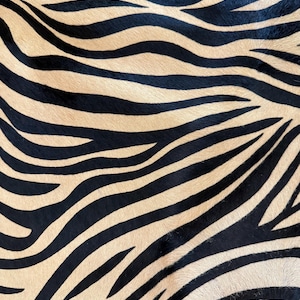 Puede incluir: Un primer plano de una alfombra de estampado de cebra con rayas negras y tostadas. La alfombra está hecha de piel de vaca y tiene una textura suave y natural.