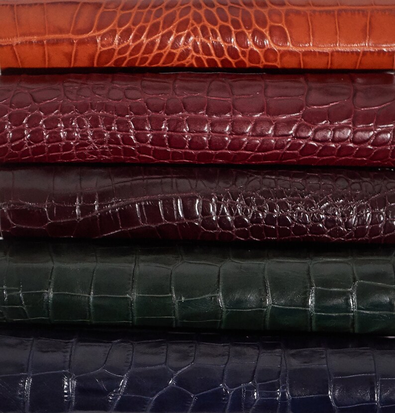 May include: Five pieces of leather stacked on top of each other. The leather is a variety of colors, including orange, burgundy, dark purple, dark green, and dark blue. The leather has a crocodile pattern.