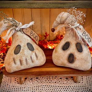 May include: Two white ghost-shaped fabric ornaments with painted faces and the word "BOO" on a tag. The ghosts are sitting on a wooden shelf with a crocheted doily underneath.