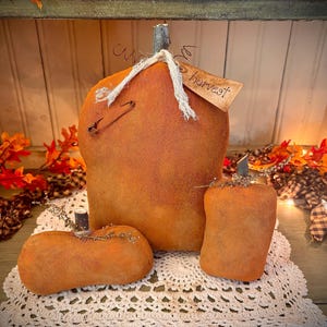 May include: Three rustic, orange, fabric pumpkins with a tag that reads "Fall Harvest". The pumpkins are sitting on a white crocheted doily.
