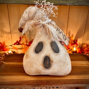 May include: A white ghost-shaped primitive doll with a black painted face and the word "BOO" on a tag. The doll is sitting on a wooden surface with autumn leaves and lights.