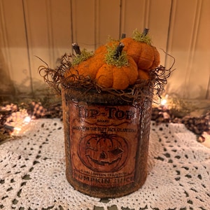 Può includere: Una lattina di metallo arrugginita con un'etichetta che recita "Tip-Top Brand Made from the Best Jack O'Lanterns A Halloween Tradition Pumpkin Juice". La lattina è piena di zucche di feltro e muschio.