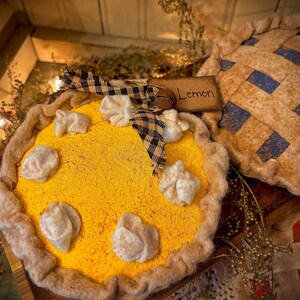 K&ouml;nnte beinhalten: Ein dekorativer Zitronenkuchen mit gelber F&uuml;llung und wei&szlig;en Akzenten, verziert mit einer karierten Schleife und einem kleinen Holzschild mit der Aufschrift "Lemon". Im Hintergrund befindet sich ein weiterer Kuchen mit blauem und beigem Gittermuster.
