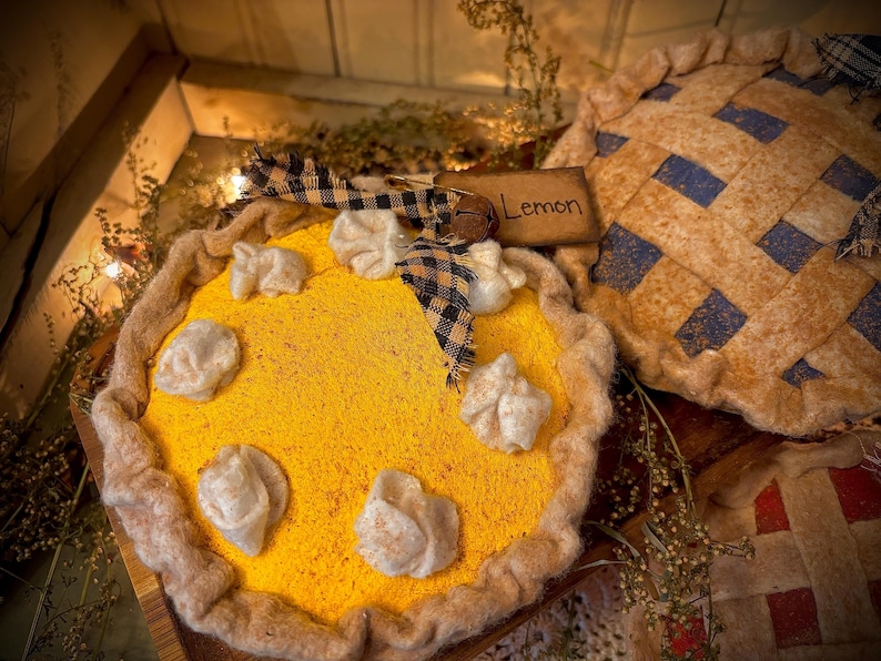 K&ouml;nnte beinhalten: Ein dekorativer Zitronenkuchen mit gelber F&uuml;llung und wei&szlig;en Akzenten, verziert mit einer karierten Schleife und einem kleinen Holzschild mit der Aufschrift "Lemon". Im Hintergrund befindet sich ein weiterer Kuchen mit blauem und beigem Gittermuster.