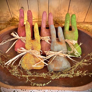 May include: Six handmade fabric bunnies in a wooden bowl. The bunnies are in various shades of pink, yellow, green, and blue. They are tied with natural raffia ribbon.