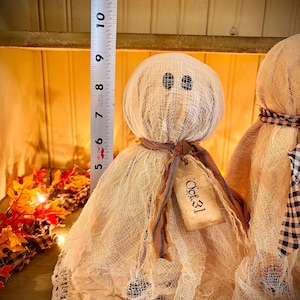 May include: Two handmade Halloween ghost dolls. The dolls are approximately 9 inches tall. Each ghost has a round head with button eyes and a draped gauze body. One ghost has a tag that reads "Oct. 31". The dolls are on a wooden surface.