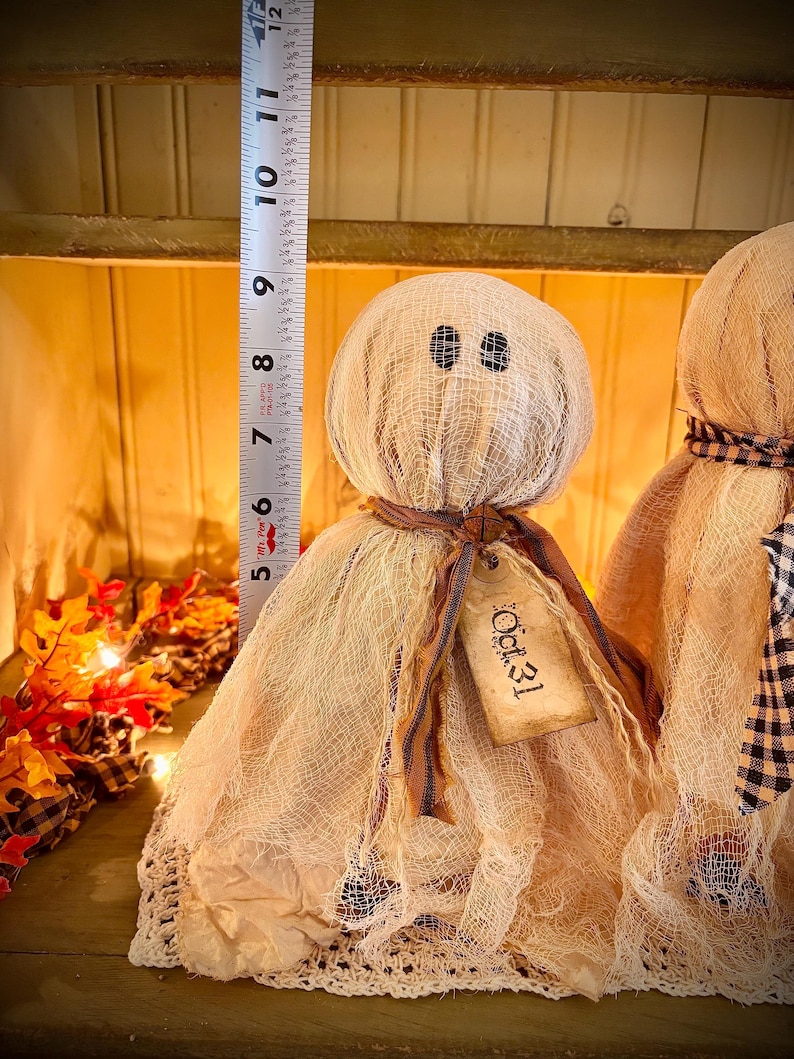 May include: Two handmade Halloween ghost dolls. The dolls are approximately 9 inches tall. Each ghost has a round head with button eyes and a draped gauze body. One ghost has a tag that reads "Oct. 31". The dolls are on a wooden surface.