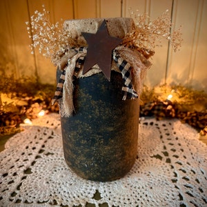 May include: A black and gold painted glass jar with a burlap and black and white checkered ribbon tied around the top. A rusty metal star sits on top of the ribbon. The jar is sitting on a white crocheted doily.