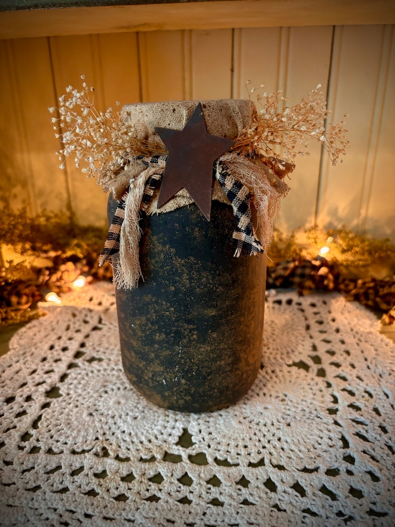 May include: A black and gold painted glass jar with a burlap and black and white checkered ribbon tied around the top. A rusty metal star sits on top of the ribbon. The jar is sitting on a white crocheted doily.