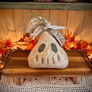 May include: A white ghost-shaped primitive doll with a black face and a tag that says "BOO" is sitting on a wooden tray with a rustic finish. The doll is decorated with dried flowers and is surrounded by autumn foliage.