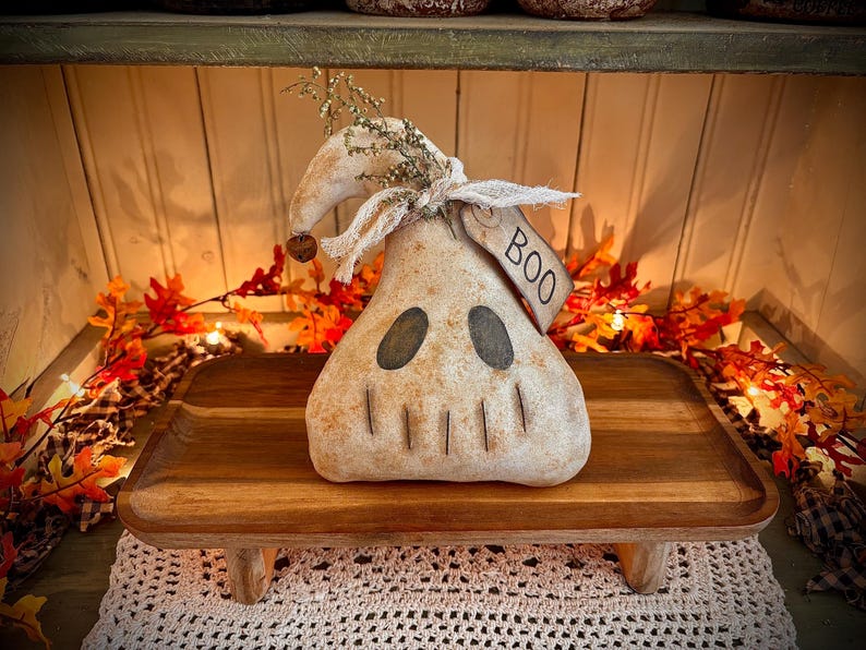 May include: A white ghost-shaped primitive doll with a black face and a tag that says "BOO" is sitting on a wooden tray with a rustic finish. The doll is decorated with dried flowers and is surrounded by autumn foliage.