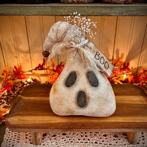 May include: A white ghost-shaped primitive doll with a black face and a burlap bow. The doll is sitting on a wooden stand with a tag that says "BOO".