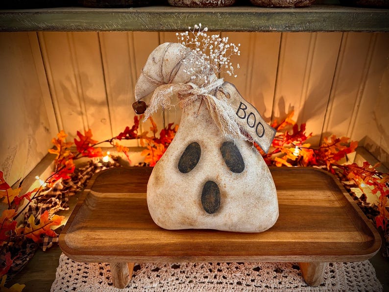 May include: A white ghost-shaped primitive doll with a black face and a burlap bow. The doll is sitting on a wooden stand with a tag that says "BOO".