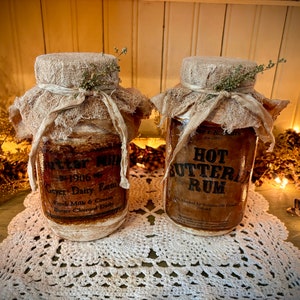 May include: Two vintage-style glass jars with fabric-covered lids. One jar reads "BUTTER MILK" and the other "HOT BUTTER RUM". Both jars have decorative labels and are tied with a ribbon and sprigs of greenery.