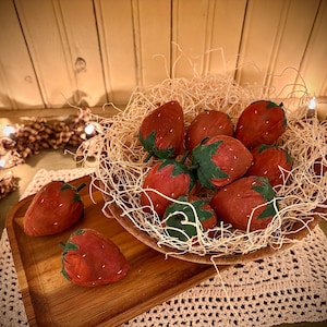 Könnte beinhalten: Eine Schüssel gefüllt mit roten Filz-Erdbeeren in einem Nest aus zerkleinertem Papier. Die Schüssel steht auf einem Holztablett mit einer weißen Häkelspitze darunter.