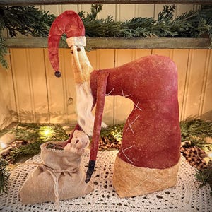 Peut inclure: Une figurine de Père Noël faite à la main, avec un long chapeau et un manteau en velours rouge. La figurine a une barbe blanche et un sac en toile de jute contenant un petit ours en peluche. Le Père Noël est posé sur un napperon en crochet blanc.