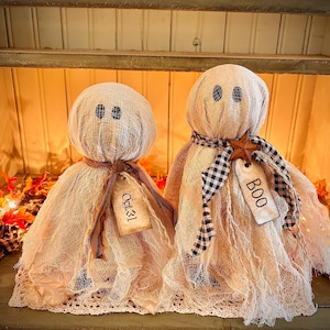 May include: Two handmade ghost decorations for Halloween. The ghosts are made of beige fabric with black button eyes and tags that say "Oct. 31" and "BOO". One ghost has a brown ribbon and the other has a black and white checkered scarf.