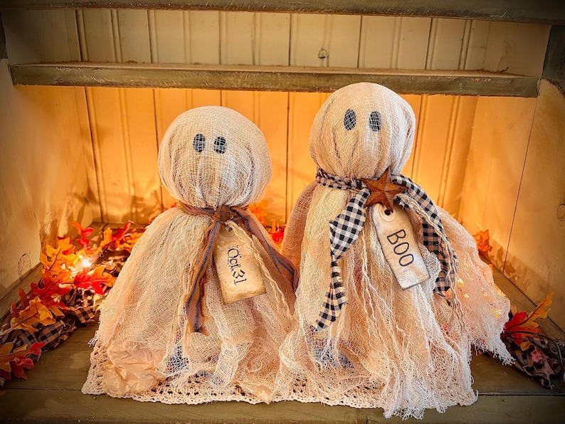 May include: Two handmade ghost decorations for Halloween. The ghosts are made of beige fabric with black button eyes and tags that say "Oct. 31" and "BOO". One ghost has a brown ribbon and the other has a black and white checkered scarf.