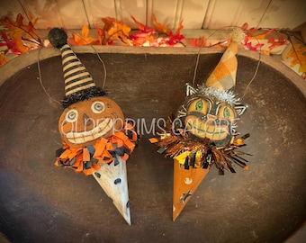 May include: Two hanging ornaments featuring a pumpkin character and a black cat character. The pumpkin wears a black and cream striped cone hat, while the cat wears an orange plaid cone hat. Both have fringed collars and wire hangers.