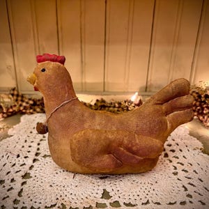 May include: A brown and red primitive style chicken figurine with a red comb and a brown and white string around its neck. The chicken is sitting on a white crocheted doily.