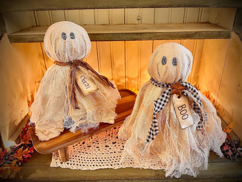 May include: Two handmade Halloween ghost decorations. Each ghost has a burlap body, stitched eyes, and a tag that says "Oct 31" or "BOO". One ghost has a brown ribbon, the other a black and white checkered ribbon.