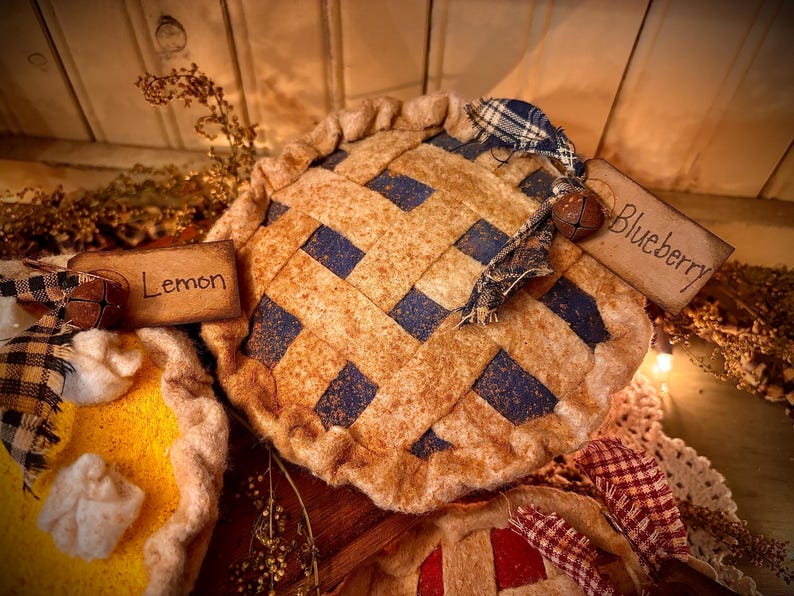 K&ouml;nnte beinhalten: Nahaufnahme von dekorativen Stoffkuchen. Ein Kuchen ist mit "Blueberry" beschriftet und hat einen Gitterteig mit blauen Quadraten. Ein weiterer Kuchen ist mit "Lemon" beschriftet und hat eine gelbe F&uuml;llung. Rustikale, l&auml;ndliche Dekoration.
