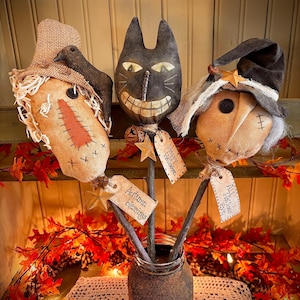 Op de afbeelding: Drie handgemaakte primitieve Halloween-figuren, een vogelverschrikker, een zwarte kat en een heks, zijn gerangschikt in een rustieke bruine pot. Elke figuur is bevestigd aan een stok met een label waarop staat "Happy Halloween", "Autumn Blessings" en "All Hallows Eve".