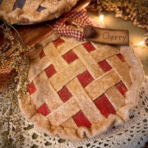 K&ouml;nnte beinhalten: Ein Filz-Kirschkuchen-Ornament mit einem Gitterkrustendesign. Der Kuchen hat eine rote F&uuml;llung und ein Holzschild mit der Aufschrift "Cherry". Eine dekorative Glocke und ein Vichy-Band sind am Schild befestigt. Das Ornament befindet sich auf einer wei&szlig;en Spitzendecke.
